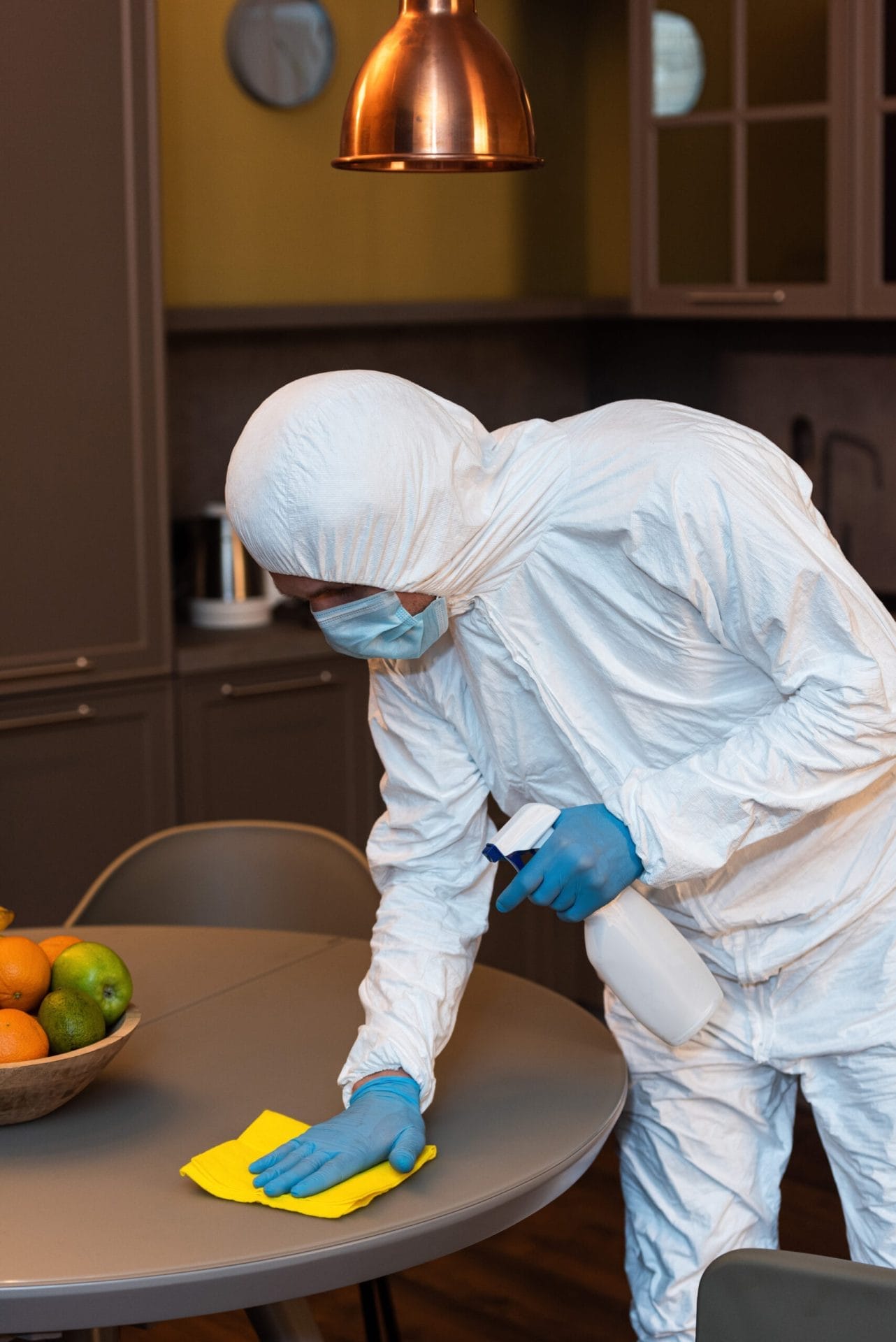 Man in hazmat suit, medical mask, and latex gloves cleaning a kitchen table with a rag and detergent, highlighting services.