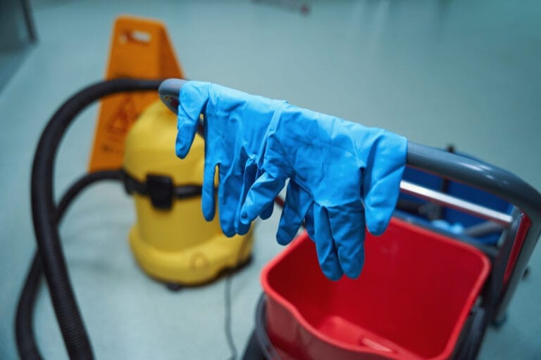 Rubber gloves on a janitor trolley next to a vacuum cleaner, emphasizing the importance of services with proper protective eq
